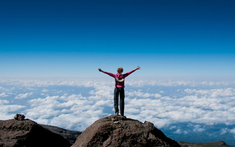 Climber with outstretched arms on the top of Mount Kilimanjaro looking down on the clouds below